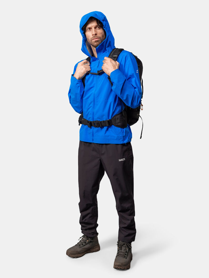 Mann in der Halti Pisarat 2,5L DX Regenjacke vor schlichtem Hintergrund, dazu schwarze Hose und Wanderschuhe.