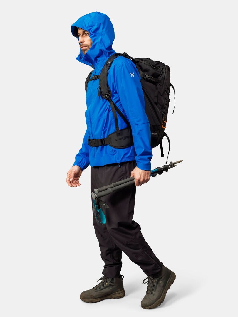 Ein Mann trägt die Halti Pisarat 2,5L DX Regenjacke, schwarze Hose, Stiefel, Rucksack und Trekkingstöcke.