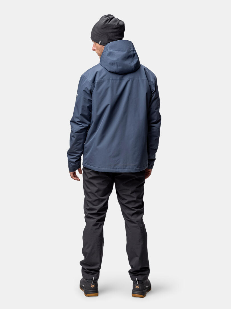 Eine Person in einer blauen Halti Forter DX Warm Regenjacke, schwarzer Hose, Stiefeln und einer grauen Mütze steht mit dem Gesicht nach außen.