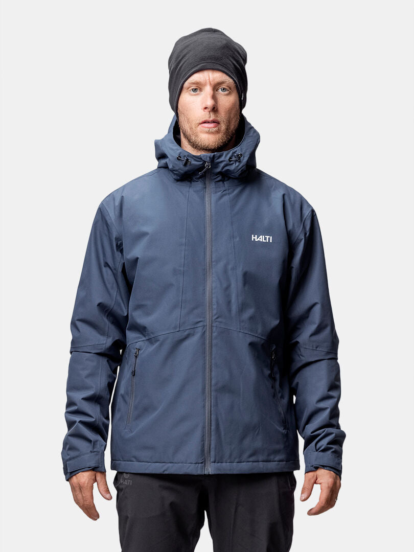 Ein Mann trägt eine Halti Forter DX Warm Regenjacke Herren und eine schwarze Mütze, mit Blick nach vorne vor einem schlichten Hintergrund.
