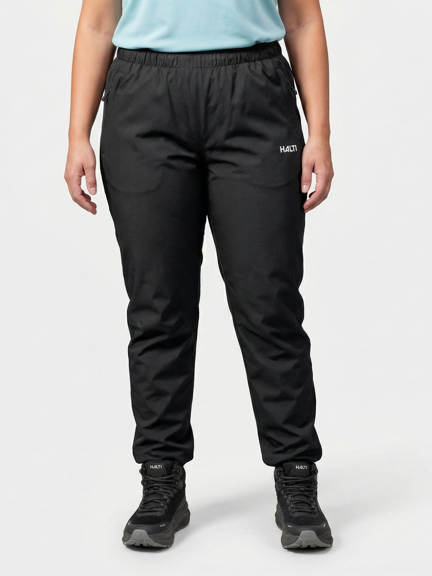 Person trägt Halti Forter DX Regenhose Plus Damen und schwarze Schuhe, stehend vor einem hellen Hintergrund, Taille nach unten.