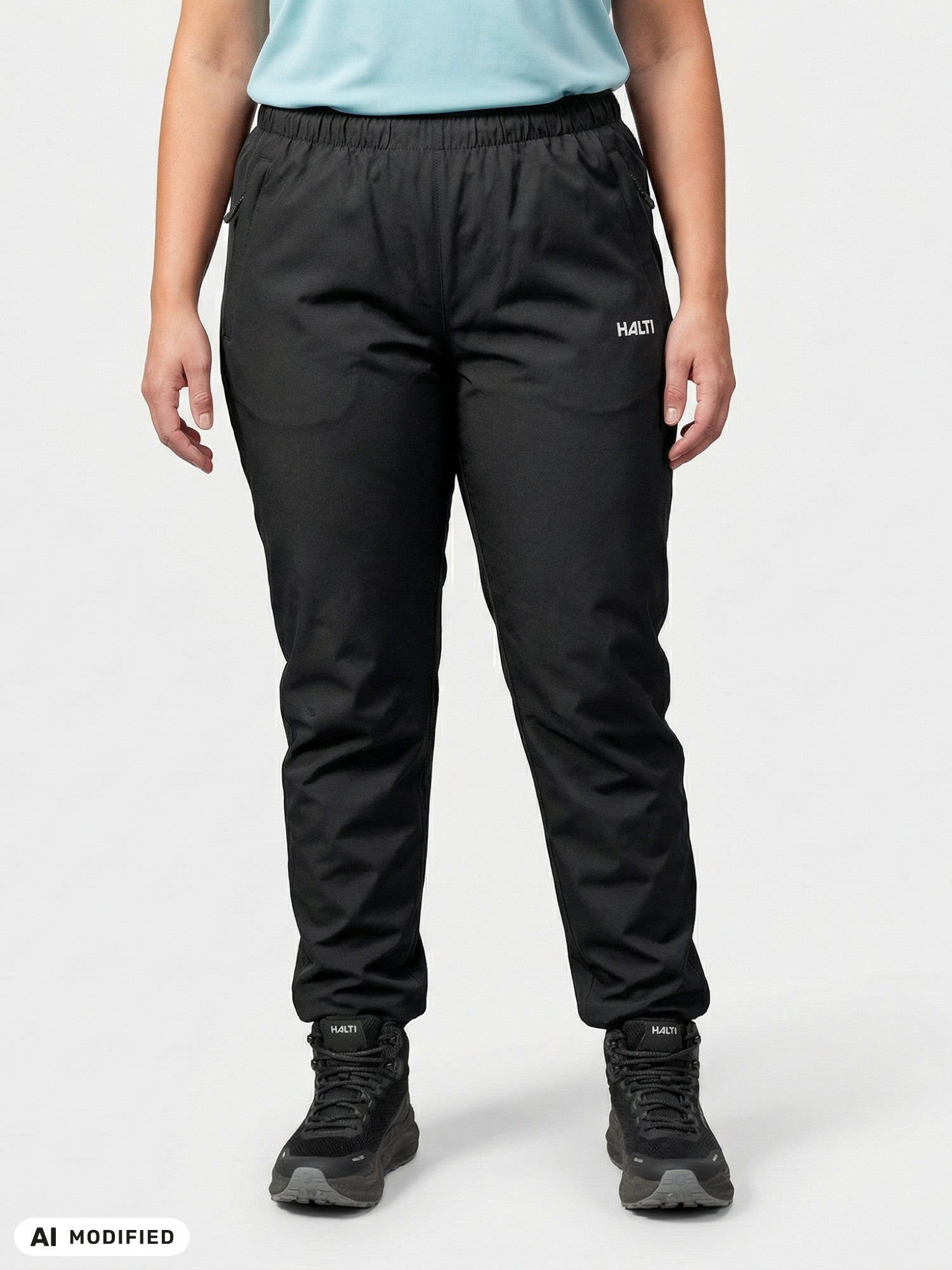 Eine Person, die eine „Halti Forter DX Regenhose Plus Damen“, schwarze Schuhe und ein hellblaues Hemd trägt, vor einem einfarbigen, hellen Hintergrund.