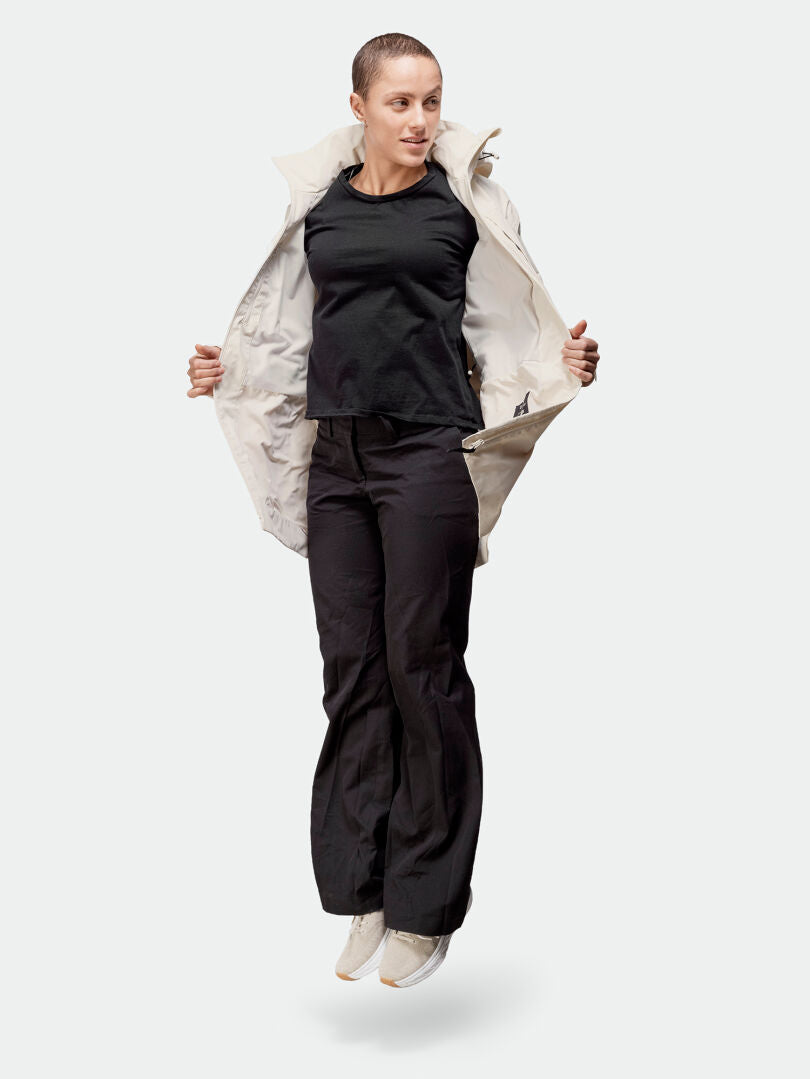 Eine Person in schwarzen Hosen, schwarzem Hemd und einer hellen Halti Kallas DX Parka Jacke Damen springt vor einem schlichten Hintergrund.