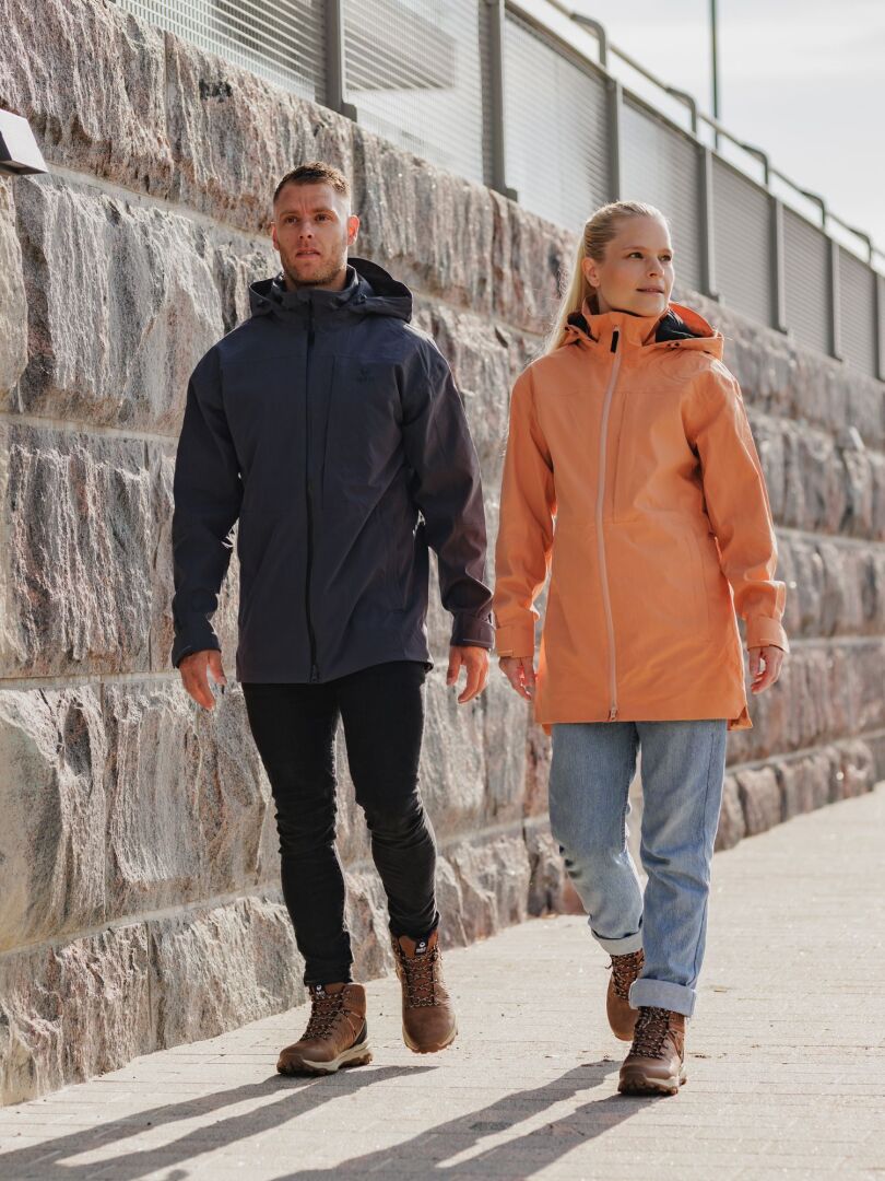 Eine Frau in einer Halti Kallas DX Parka Jacke für Damen und Stiefeln geht an einem sonnigen Tag an einer Steinmauer entlang.