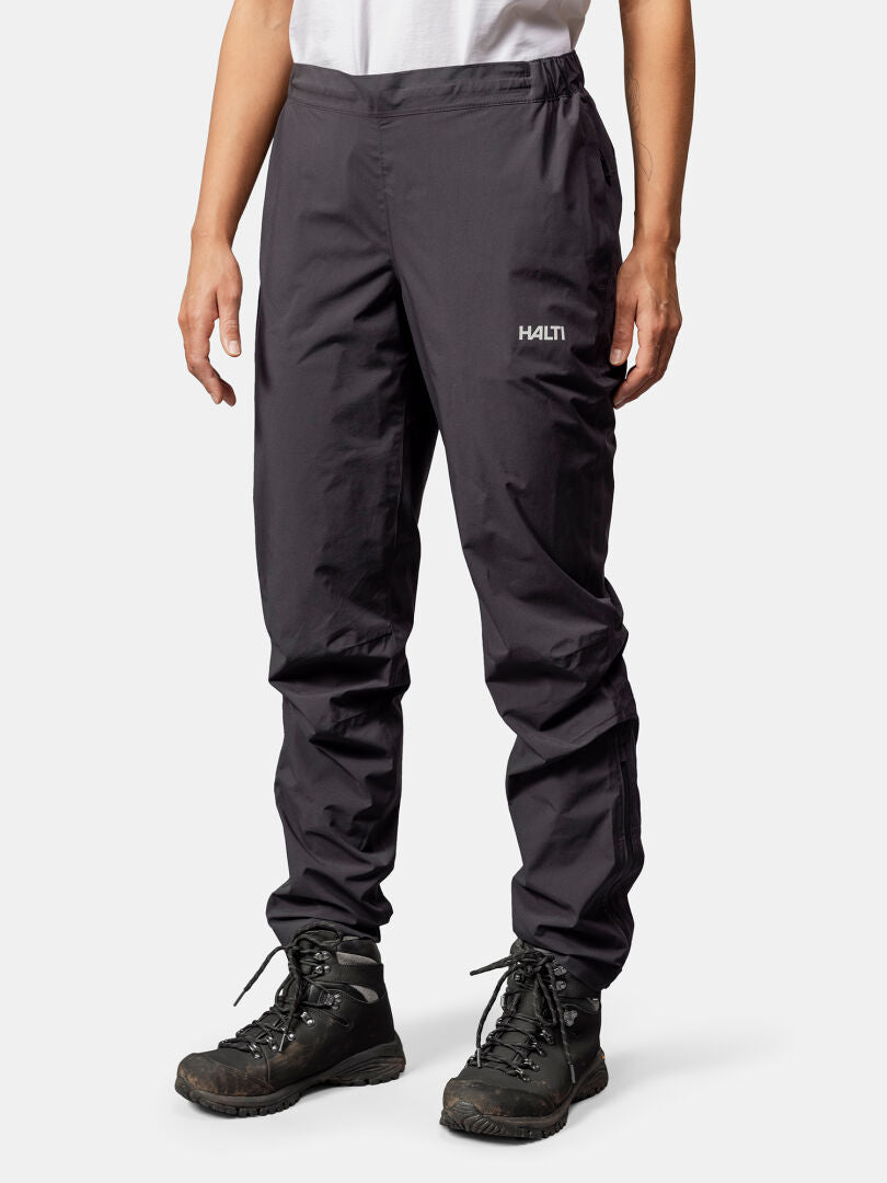 Eine Frau trägt Halti Pisarat 2,5L DX Regenhosen, schwarze Wanderschuhe und ein weißes Shirt vor neutralem Hintergrund.