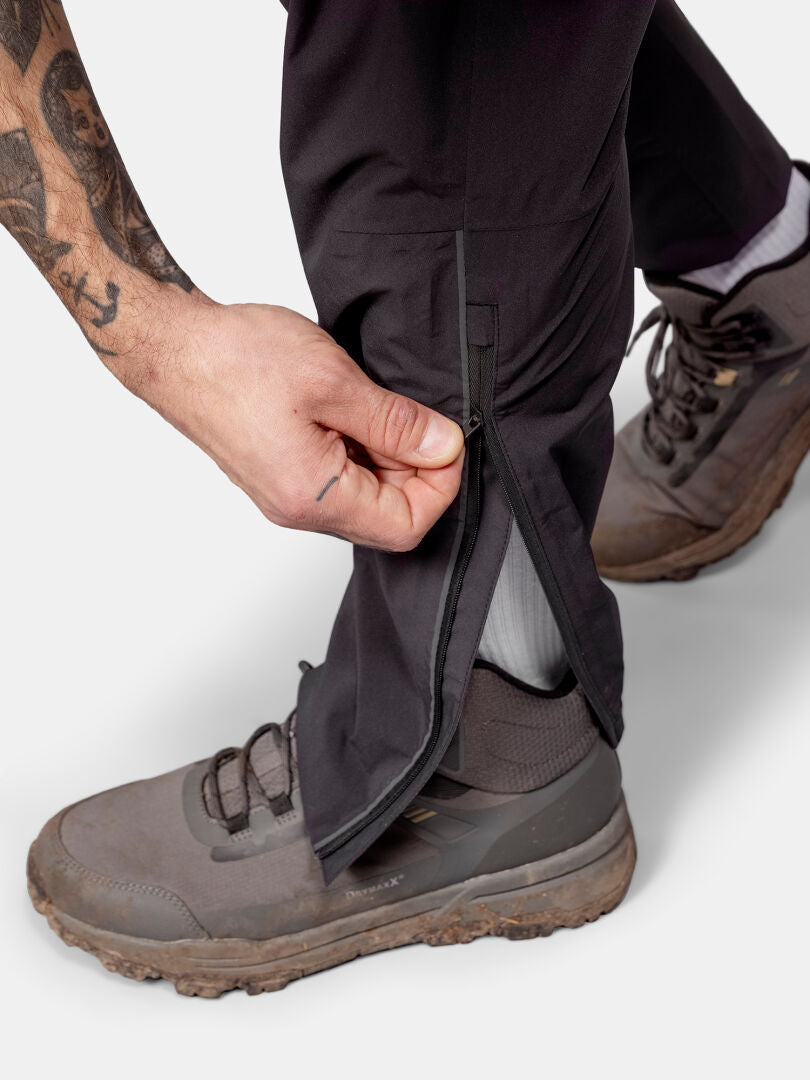 Eine Person öffnet die Manschette der Halti Pisarat 2,5L DX Regenhosen Herren und trägt graue Wanderschuhe.