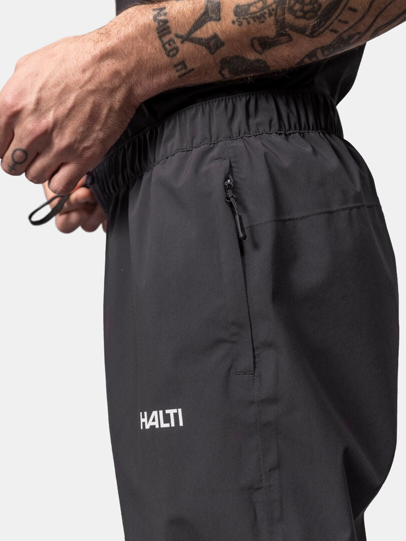 Eine Person stellt den Kordelzug der schwarzen Halti Pisarat 2,5L DX Regenhosen Herren ein und zeigt dabei tätowierte Unterarme.