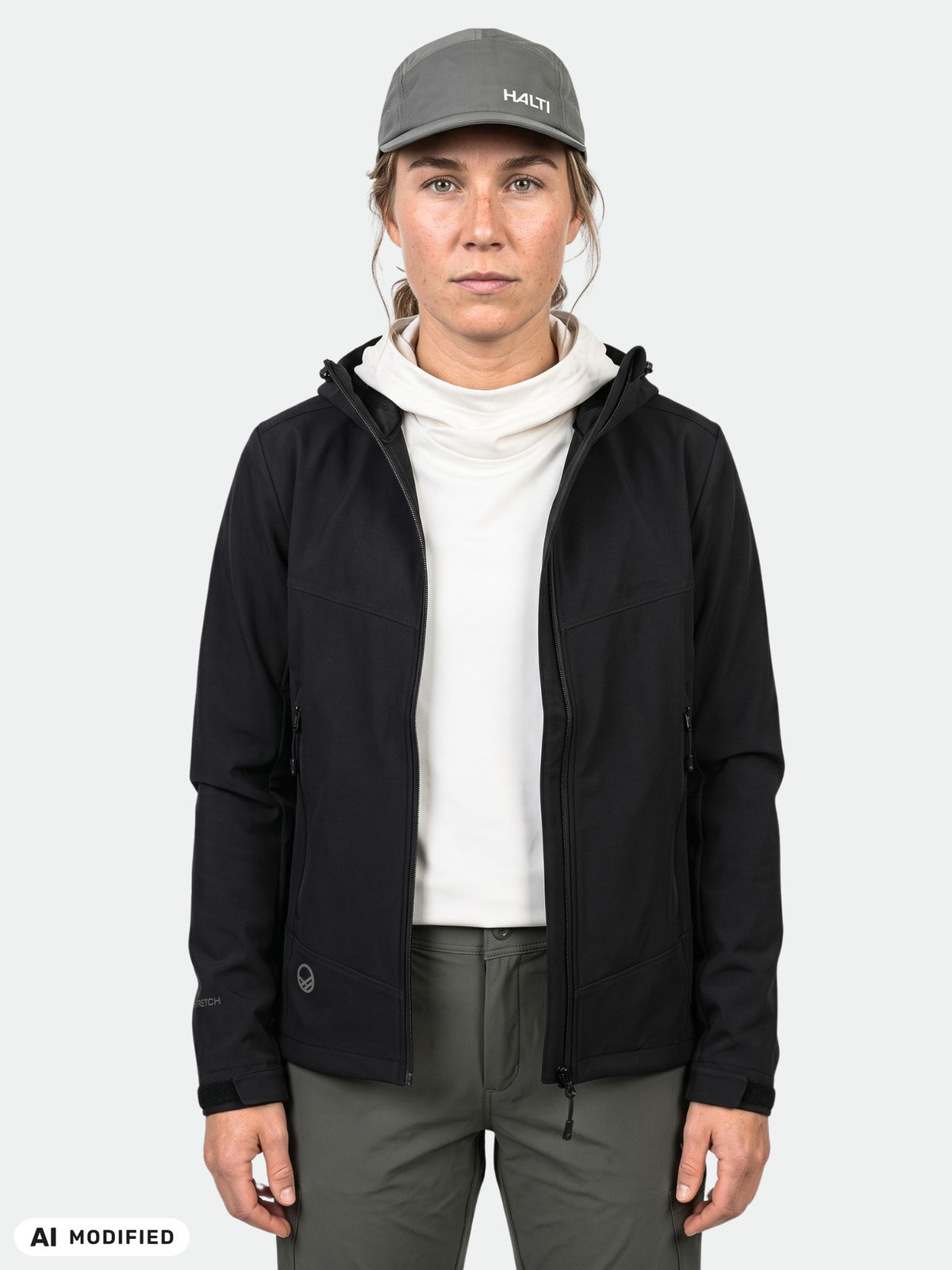Eine Person in einer schwarzen Halti Pallas Evo Brushed X-Stretch-Jacke für Damen, einem weißen Oberteil mit hohem Kragen, einer grauen Hose und einer grauen Mütze.