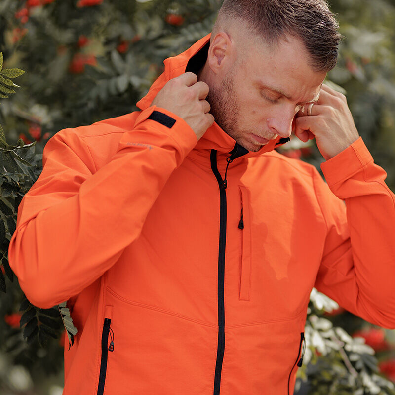 Ein Mann richtet die Kapuze seiner Halti Pallas Evo Brushed X-Stretch Jacke für Herren im Freien in der Nähe von grünem Laub und roten Beeren.