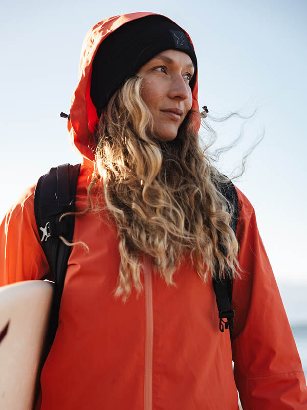 Person mit langen gewellten Haaren in einer Halti Staala DX Regenjacke Damen und schwarzer Mütze hält ein Surfbrett im Freien.