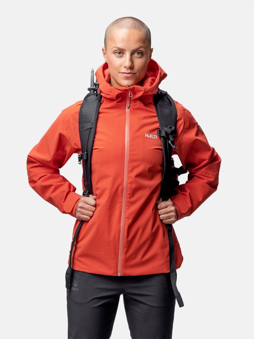 Eine Frau trägt eine rote Halti Staala DX Regenjacke und einen schwarzen Rucksack und blickt in die Kamera vor einem schlichten Hintergrund.