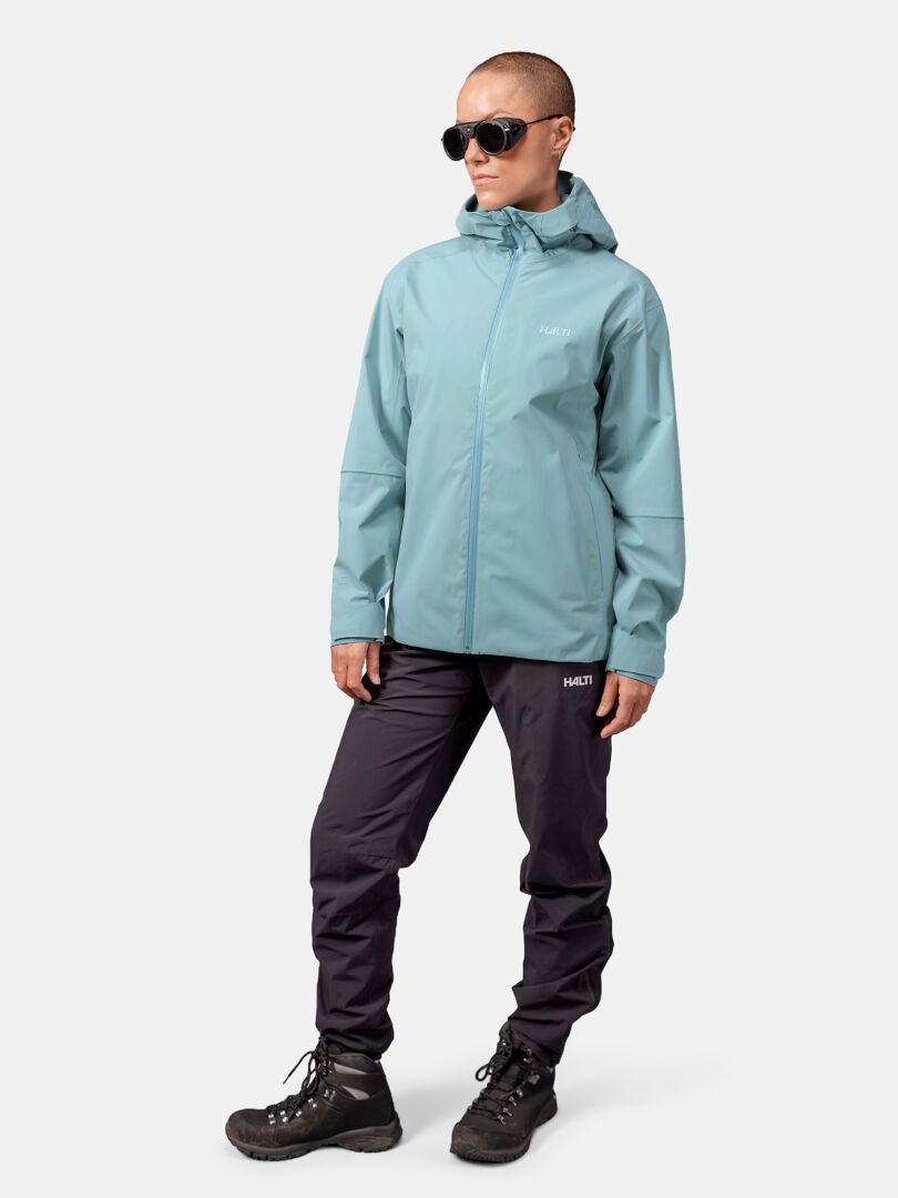 Eine Person in einer hellblauen Halti Staala DX Regenjacke Damen, einer schwarzen Hose und einer Sonnenbrille steht vor einem schlichten Hintergrund.