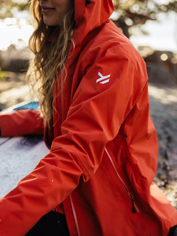 Eine Person mit langen Haaren trägt die rote Halti Staala DX Regenjacke Damen mit weißem Logo neben einem Surfbrett im Freien.