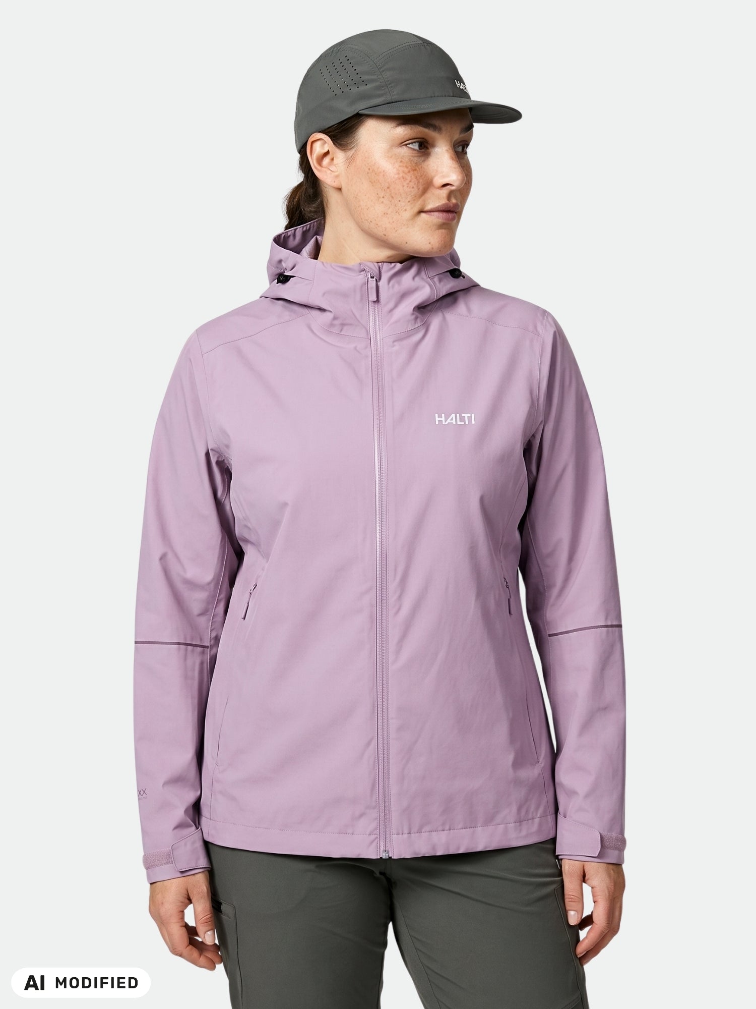 Eine Person in einer hellvioletten Halti Staala DX Regenjacke Damen Plus und einer grauen Mütze, die vor einem schlichten, hellen Hintergrund steht.
