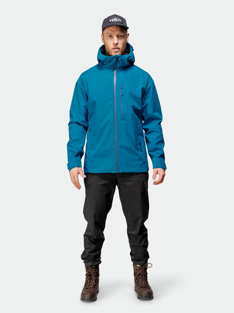Ein Mann in einer blauen Halti Staala DX Regenjacke Herren, einer schwarzen Hose, braunen Stiefeln und einer schwarzen Mütze steht vor uns.