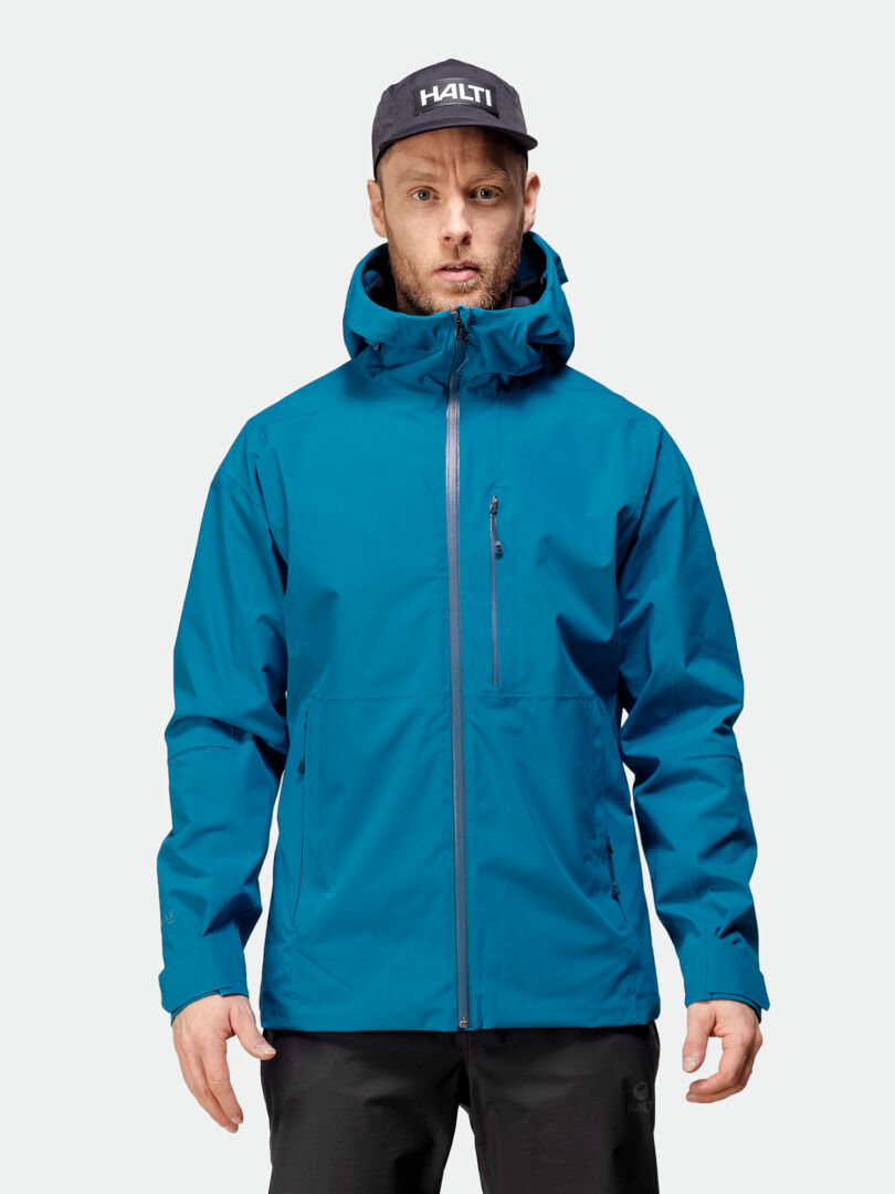 Ein Mann trägt eine blaue Halti Staala DX Regenjacke Herren und eine schwarze Hose, bereit für Abenteuer im Freien vor einem schlichten Hintergrund.
