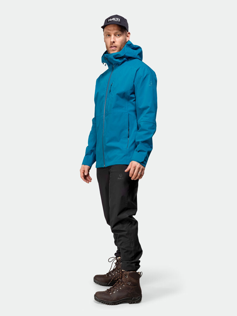 Ein Mann in einer blauen Halti Staala DX Regenjacke, schwarzer Hose und braunen Wanderschuhen steht bereit für Outdoor Abenteuer.