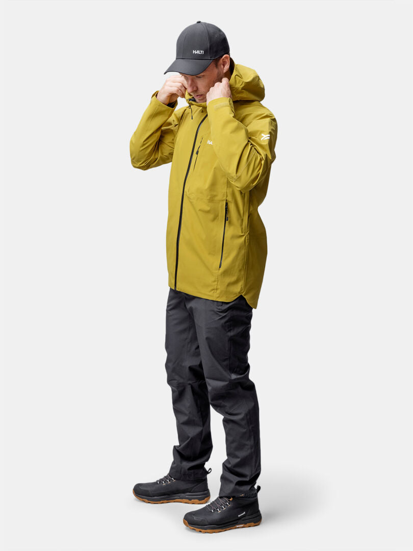 Eine Person rückt die Kapuze ihrer Halti Staala DX Regenjacke Herren zurecht, einer gelben Jacke, bereit für ein Outdoor-Abenteuer.