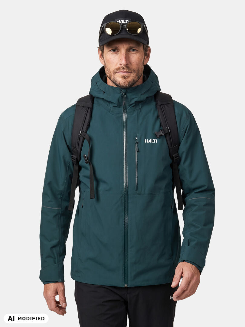 Ein Mann in einer blaugrünen Halti Staala DX Regenjacke für Herren mit Kapuze, einem schwarzen Rucksack und einer Mütze steht mit dem Blick nach vorne.