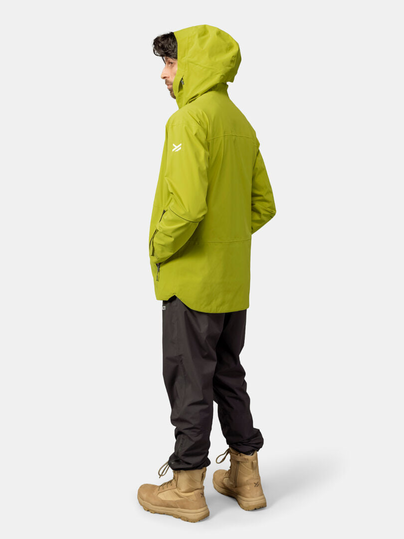 Mann in grüner Halti Staala DX Regenjacke, schwarzer Hose und hellbraunen Stiefeln steht seitlich vor einem schlichten hellen Hintergrund.
