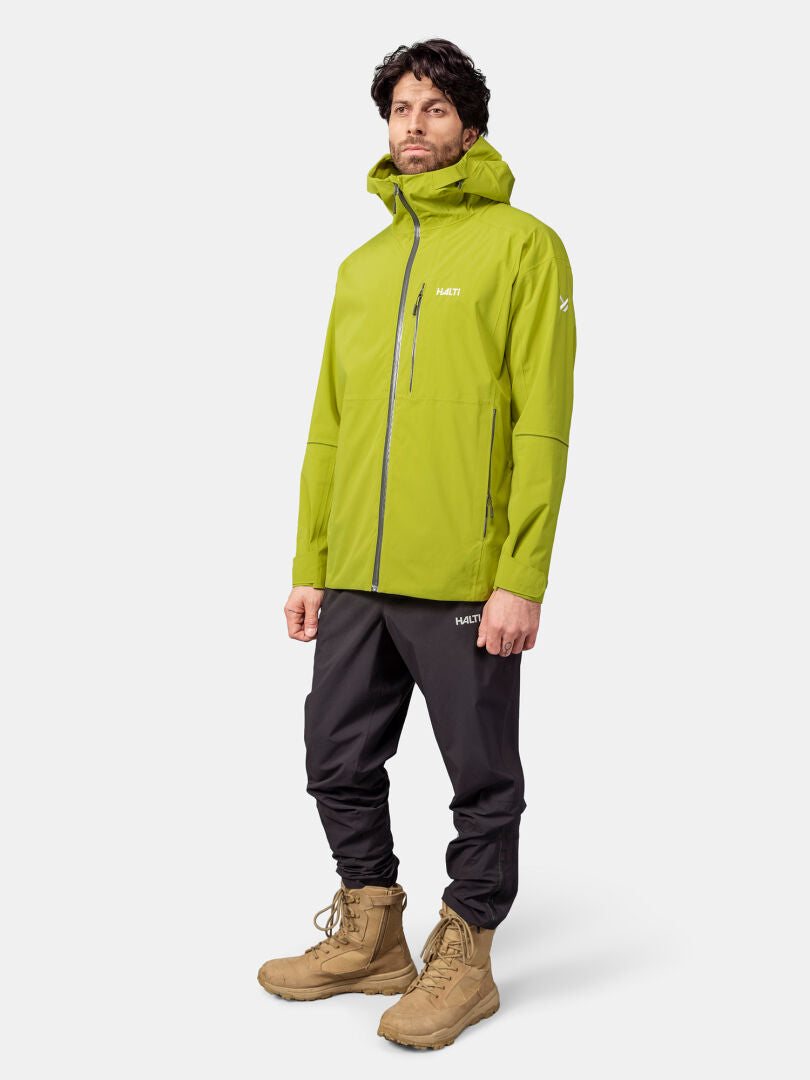 Ein Mann trägt eine lindgrüne Halti Staala DX Regenjacke Herren mit schwarzen Hosen und hellbraunen Stiefeln vor einem weißen Hintergrund.