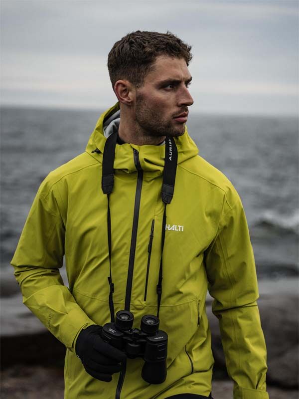 Ein Mann in einer Halti Staala DX Regenjacke Herren mit Fernglas steht am Meer, bereit für Abenteuer.
