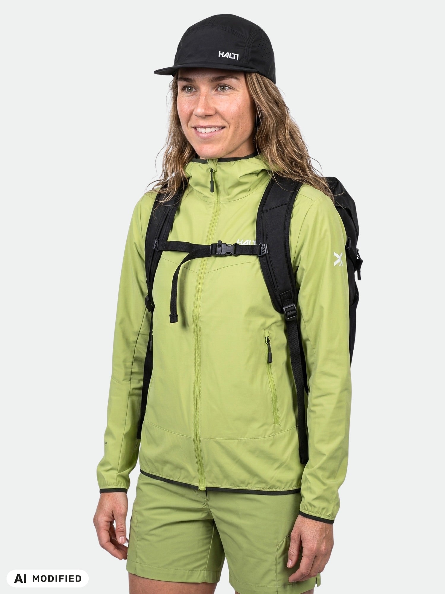 Frau in einer Halti Pallas Cool X-Stretch-Jacke und Shorts, mit schwarzer Mütze und Rucksack vor einem neutralen Hintergrund.