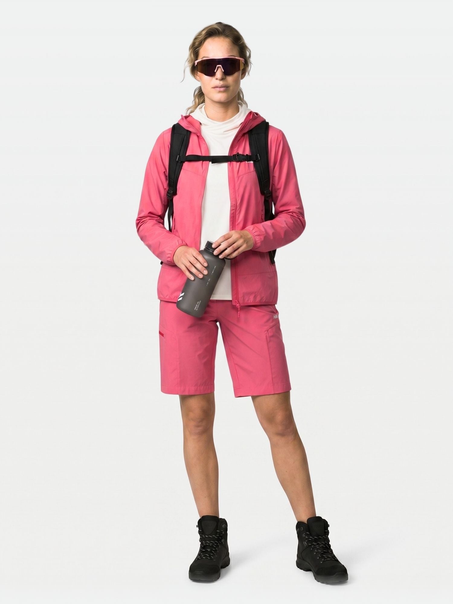 Eine Person in einer rosafarbenen Halti Pallas Cool X-stretch Jacke Damen, Shorts, schwarzen Stiefeln, Sonnenbrille, Rucksack, hält einen Becher in der Hand.