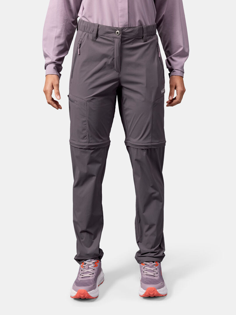 Eine Frau in Halti Pallas Cool Stretch Zip-off Hosen, einem langärmeligen Shirt und lila Sneakers steht vor einem weißen Hintergrund.