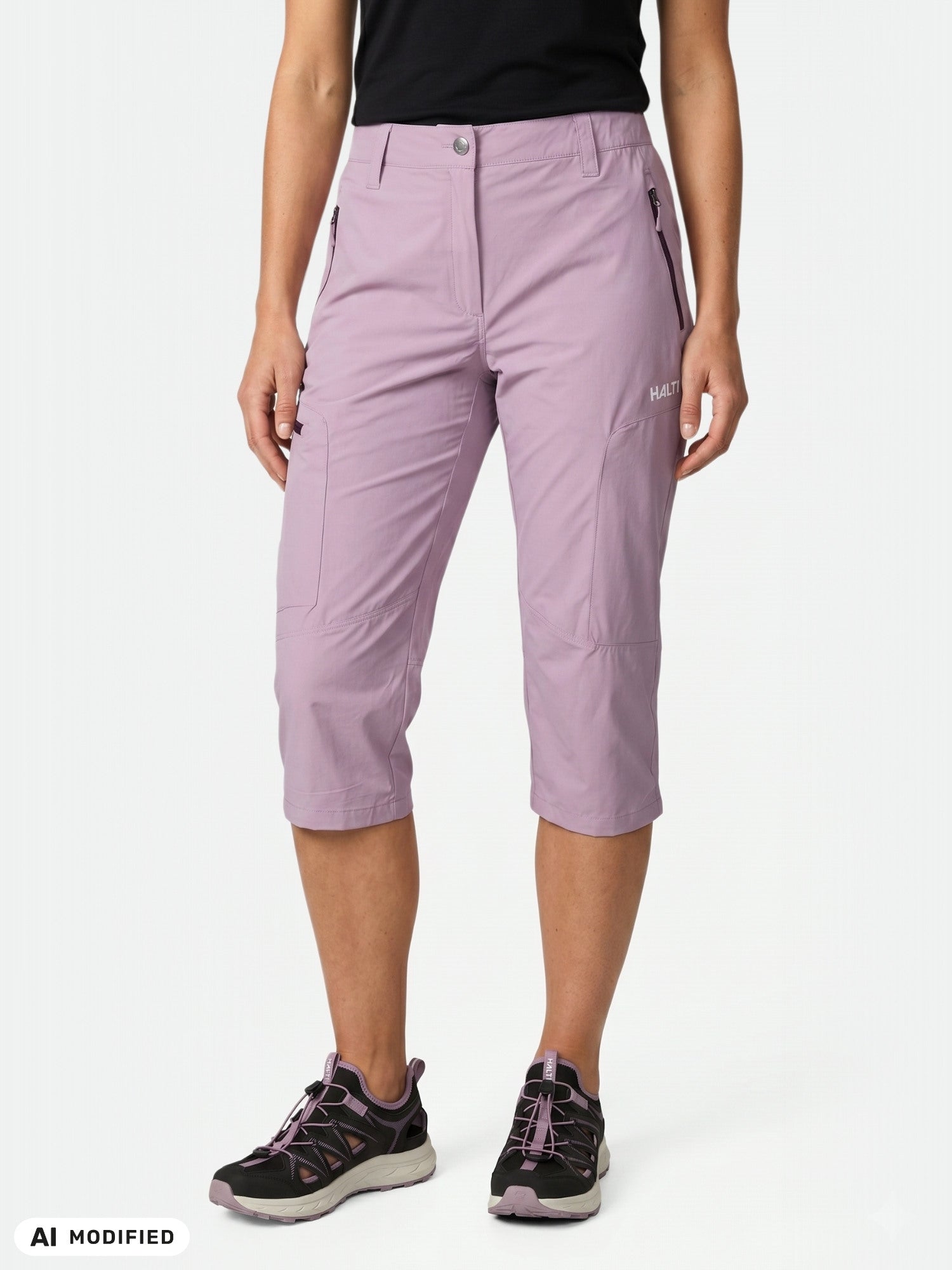 Frau in einer „Halti Pallas Cool X-Stretch“-Caprihose für Damen, schwarzem T-Shirt und Sneakers vor weißem Hintergrund.