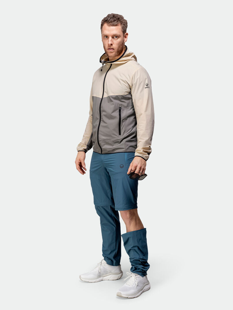 Ein Mann trägt die Halti Pallas Cool X-Stretch Jacke Herren, eine blaue Zip-Off-Hose und weiße Sneakers vor einem schlichten Hintergrund.