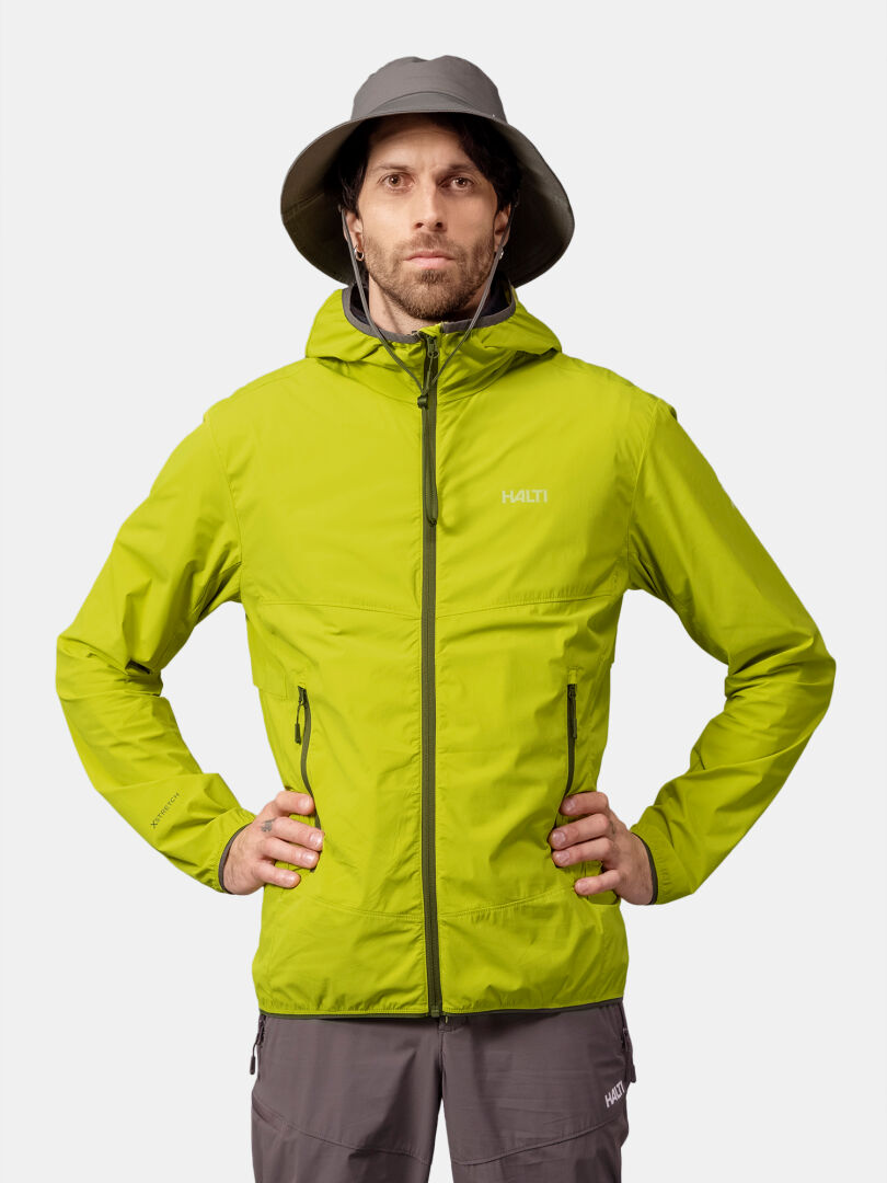 Ein Mann in einer grünen Halti Pallas Cool X-stretch Herrenjacke und grauem Hut steht mit den Händen in den Hüften vor einem schlichten Hintergrund.