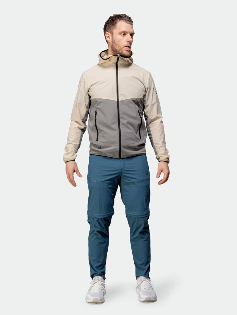 Ein Mann trägt die Halti Pallas Cool Stretch Zip-off Hose in Blau, eine beige-graue Kapuzenjacke und weiße Turnschuhe.
