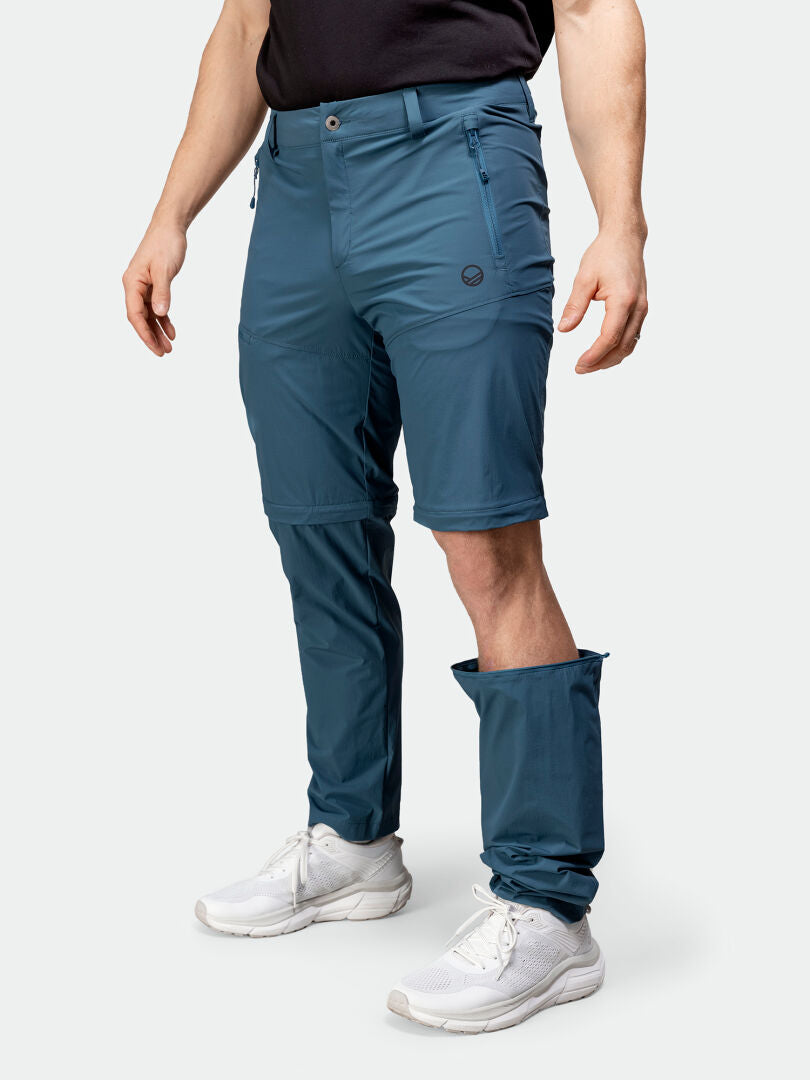 Eine Person in Halti Pallas Cool Stretch Zip-off Hosen Herren, einem schwarzen Hemd und weißen Turnschuhen steht vor einem weißen Hintergrund.
