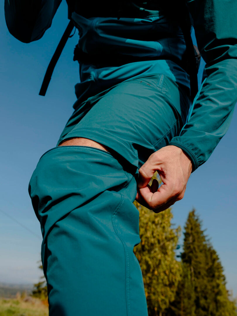 Eine Person zieht im Freien mit Bäumen und blauem Himmel den unteren Teil der Halti Pallas Cool X-Stretch Zip-Off-Hose für Herren ab.