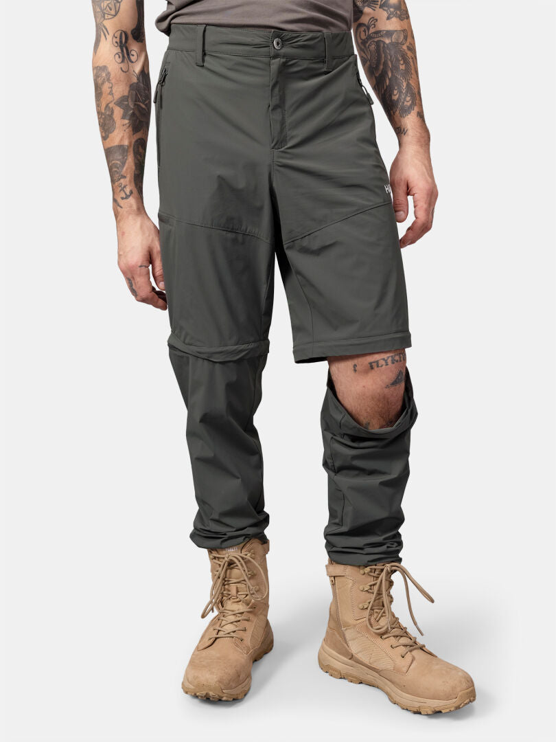 Eine Person in Halti Pallas Cool Stretch Zip-off Hosen Herren, mit aufgeschlagenen Knien und hellbraunen Stiefeln auf weißem Hintergrund.