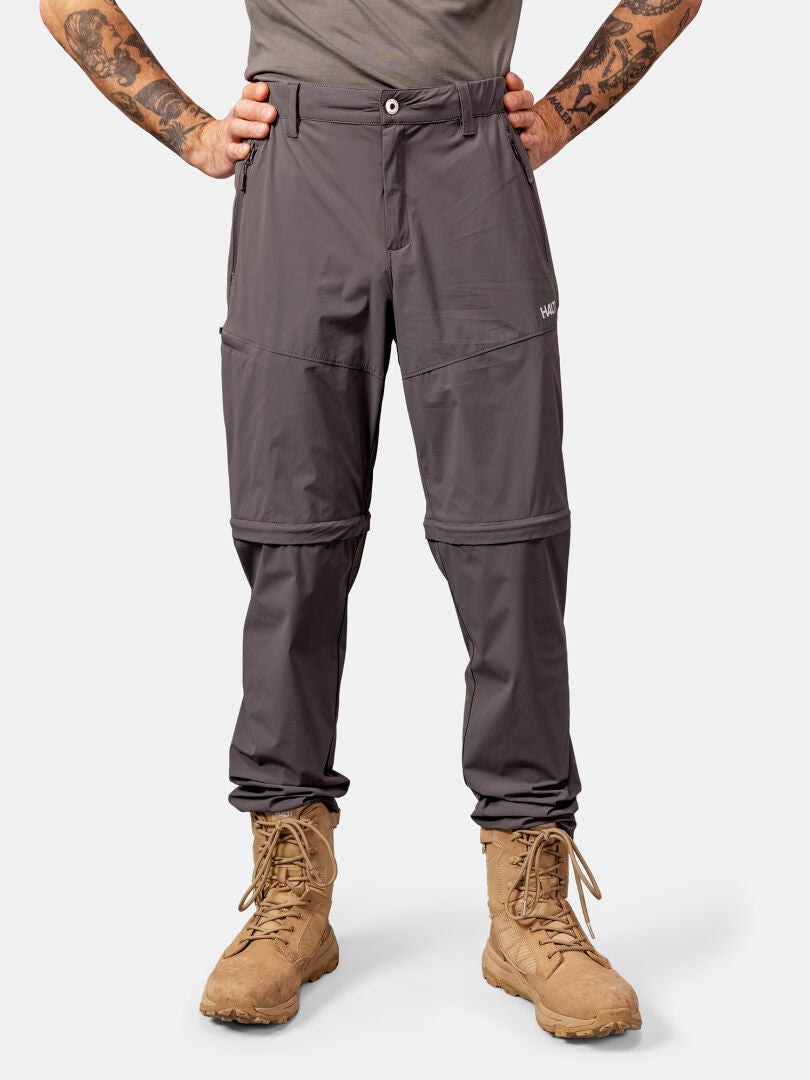 Mann in Halti Pallas Cool Stretch Zip-off Hosen und braunen Schnürstiefeln, stehend, Hände in die Hüften gestemmt, vor einem schlichten Hintergrund.