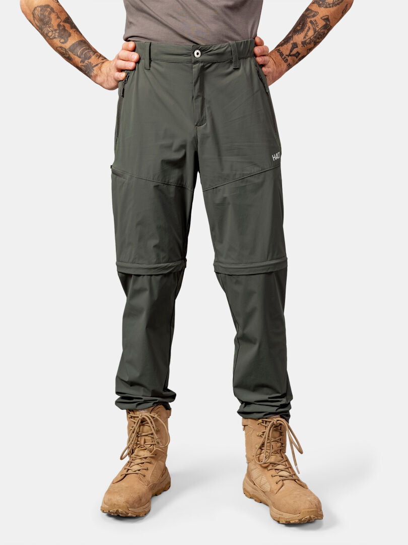 Mann in Halti Pallas Cool Stretch Zip-off Hosen und braunen Schnürstiefeln, stehend, Hände in die Hüften gestemmt, vor einem schlichten Hintergrund.