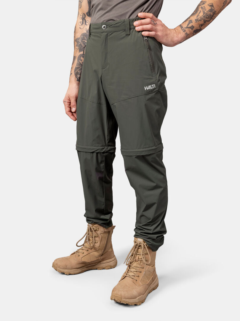 Eine Person trägt Halti Pallas Cool Stretch Zip-off Hosen Herren und ein graues Hemd und posiert mit einer Hand in der Hüfte.