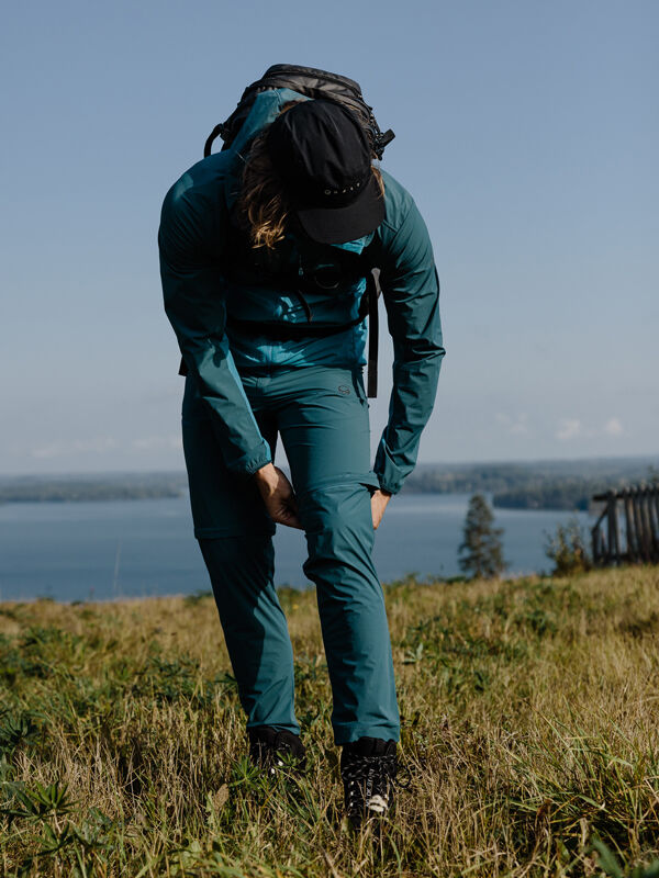 Eine Person in der Halti Pallas Cool X-Stretch Zip-Off-Hose für Herren richtet ihr Hosenbein, während sie auf dem Gras am Wasser steht.
