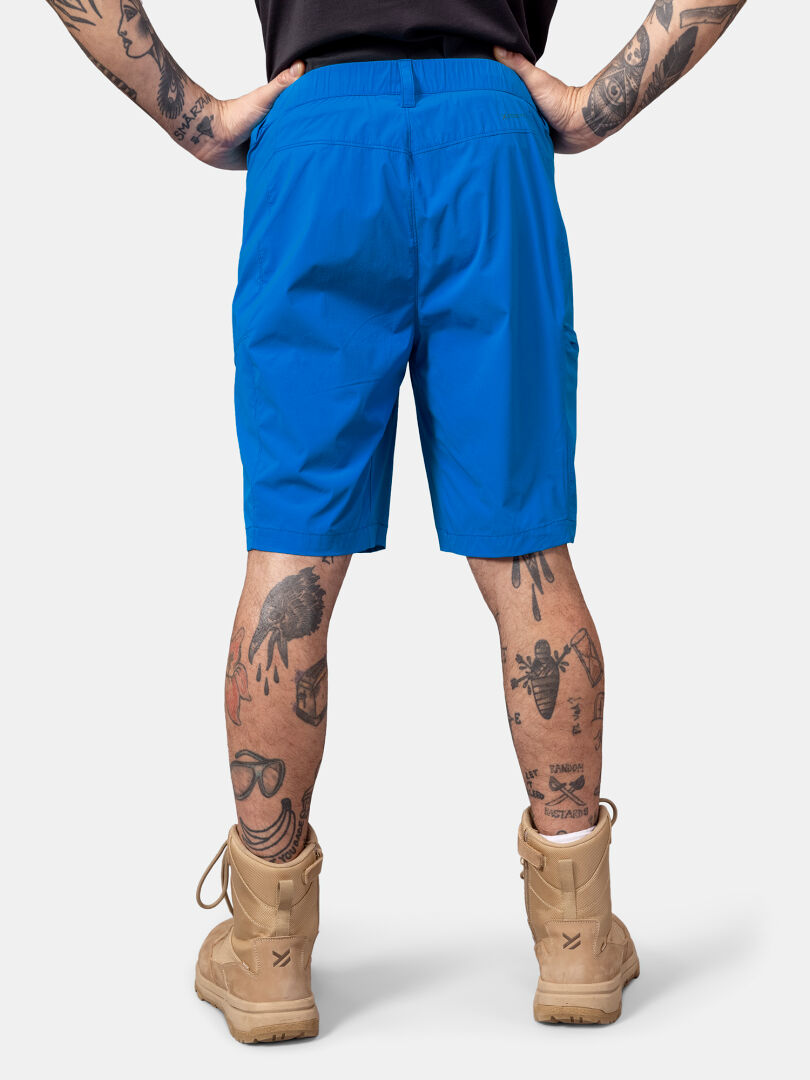 Person in hellblauen Halti Pallas Cool X-stretch Shorts, hellbraunen Kampfstiefeln, schwarzem Hemd, mit Tätowierungen an Armen und Beinen.