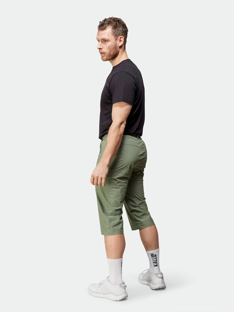 Ein Mann steht seitlich in einem schwarzen T-Shirt, Halti Pallas Cool X-Stretch Caprihose Herren, weißen Socken und Turnschuhen.