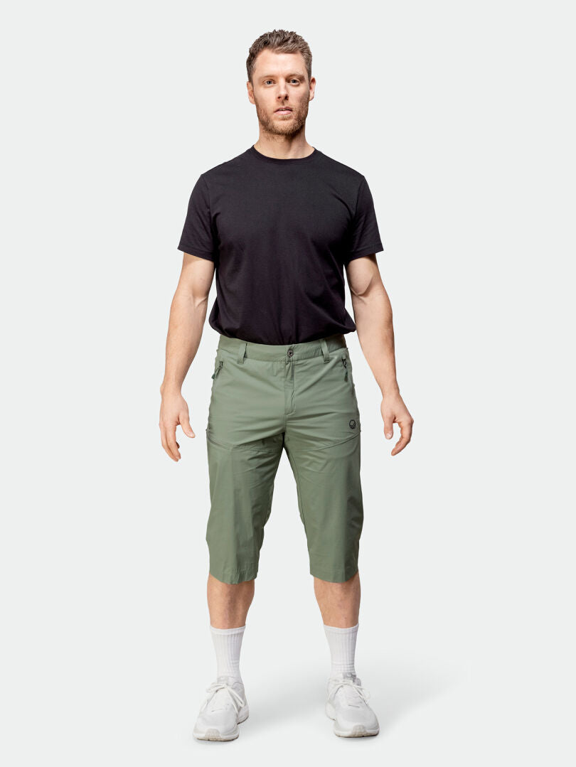 Ein Mann trägt Halti Pallas Cool X-Stretch Caprihose Herren mit einem schwarzen T-Shirt, weißen Socken und Turnschuhen.