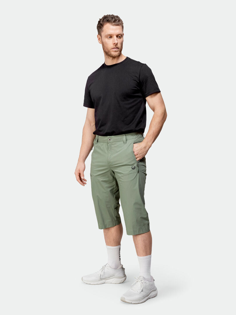 Der Mann trägt ein schwarzes T-Shirt, Halti Pallas Cool X-Stretch Caprihose Herren, weiße Socken und Turnschuhe vor einem schlichten Hintergrund.