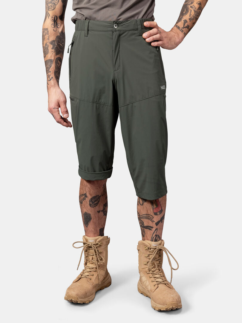 Aufrecht stehende Person in Halti Pallas Cool X-stretch Caprihose Herren, taupefarbenem T-Shirt, hellbraunen Stiefeln, Tattoos an Armen und Beinen.