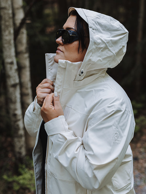 Eine Frau in einer hellen Kallas DX Parka Jacke Plus Damen von Halti und mit Sonnenbrille steht im Freien und hält den Kragen.