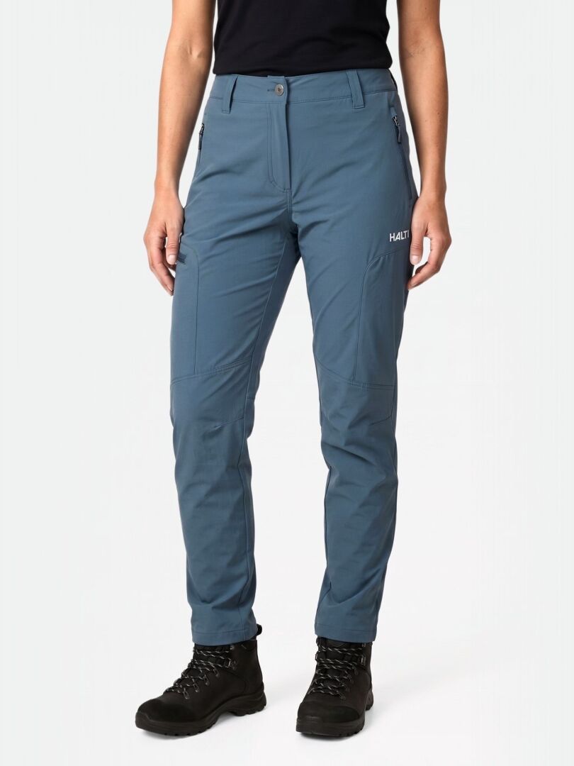 Eine Person trägt Halti Pallas Cool X-stretch Hosen Damen, schwarze Stiefel und ein Hemd vor einem schlichten hellen Hintergrund.