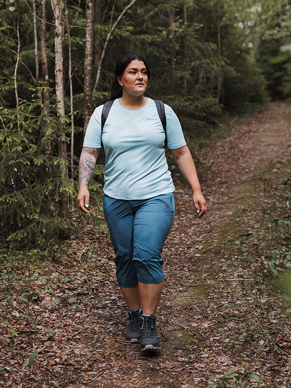 Person mit dunklem Haar und Rucksack geht in Halti Pallas Cool X-Stretch Caprihose Damen Plus und hellblauem Hemd auf einem Waldweg.