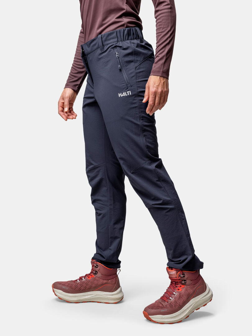 Eine Person in Halti Finholma X-stretch Hose Damen, einem langärmeligen Hemd und Wanderschuhen steht vor einem weißen Hintergrund.