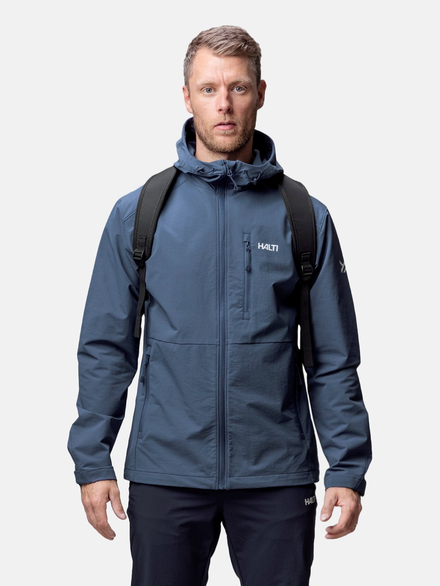 Ein Mann in einer Halti Finholma X-Stretch Jacke Herren und einem schwarzen Rucksack steht mit dem Gesicht nach vorne vor einem schlichten hellen Hintergrund.